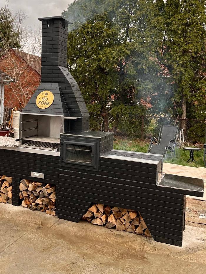 Estação BBQ Zone preta com churrasqueira em aço inox, forno com porta de ferro, chapa de cocção e bancada de granito.