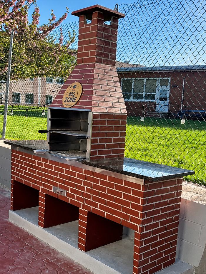 Churrasqueira BBQ Zone de tijolo vermelho com chaminé, grelhas em aço inox e bancada de granito, instalada sobre base de concreto.
