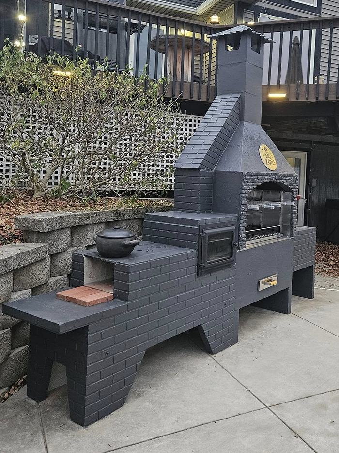 Churrasqueira BBQ Zone cinza escura com fogão a lenha integrado, chapa de cocção com panela de barro e bancada lateral.