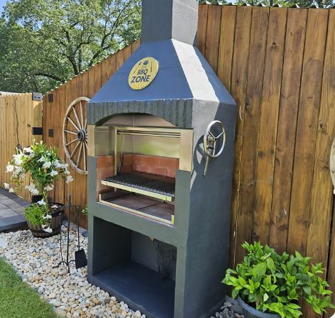 Churrasqueira BBQ Zone azul acinzentada em um canto de jardim, com cerca de madeira, plantas e pedras decorativas.