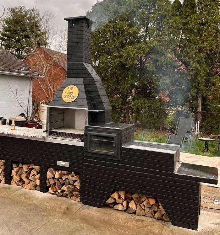 Churrasqueira BBQ Zone preta com forno fechado, bancada de granito e lenha organizada em nichos sob a estrutura, instalada em um quintal.
