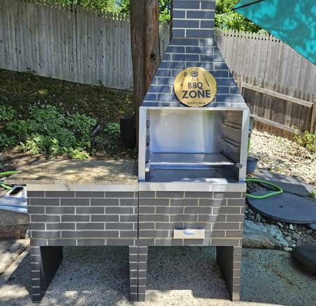 Churrasqueira BBQ Zone com acabamento de tijolo cinza e bancada, recém-instalada em um quintal com cerca de madeira.