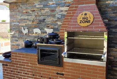 Churrasqueira BBQ Zone de tijolo vermelho com fogão a lenha e panelas de barro, integrada a uma área externa com parede de pedra.