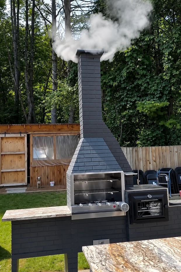 Churrasqueira BBQ Zone escura em funcionamento, com fumaça saindo da chaminé, grelhas reguláveis e forno lateral com porta de vidro.